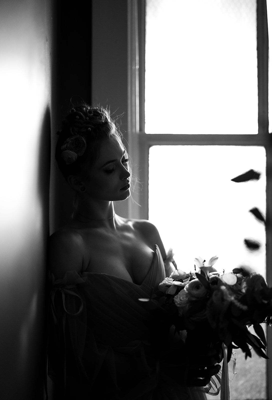 A black and white photo of a woman leaning against a wall holding a bouquet of flowers.