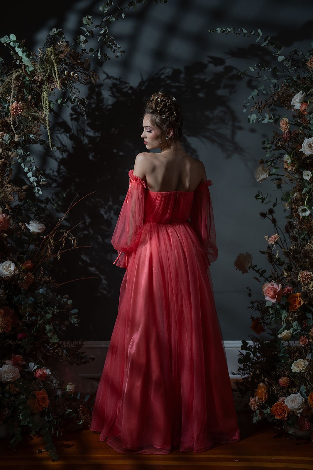 A woman in a red dress is standing in front of a wall of flowers.