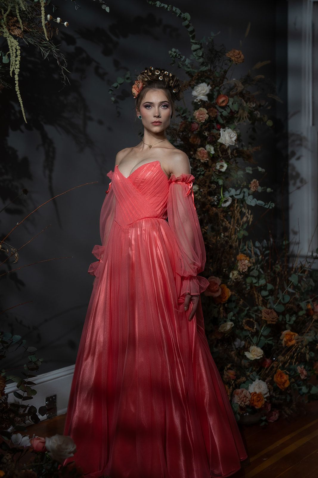 A woman in a red dress is standing in front of a wall of flowers.