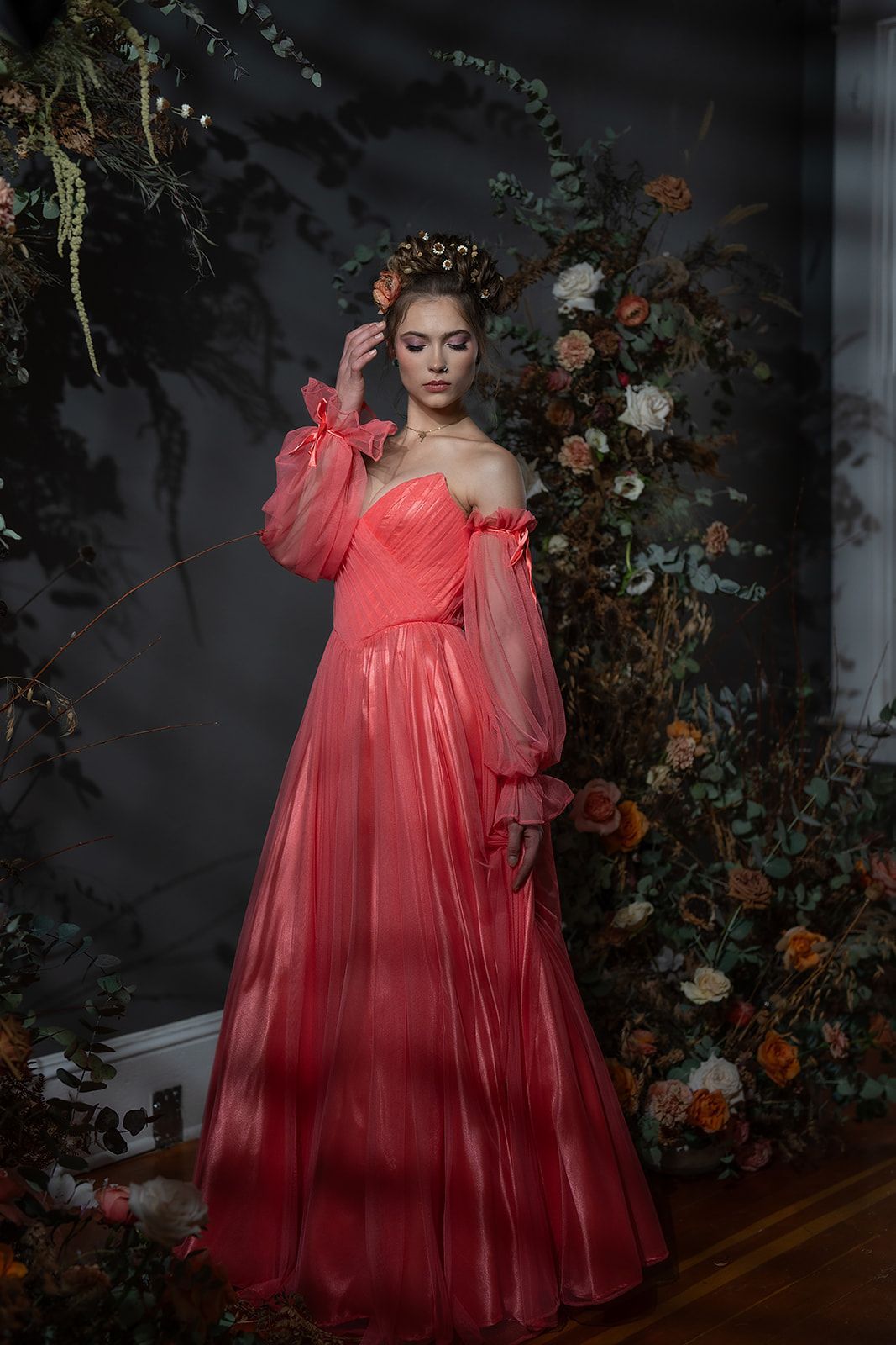 A woman in a red dress is standing in front of a wall of flowers.