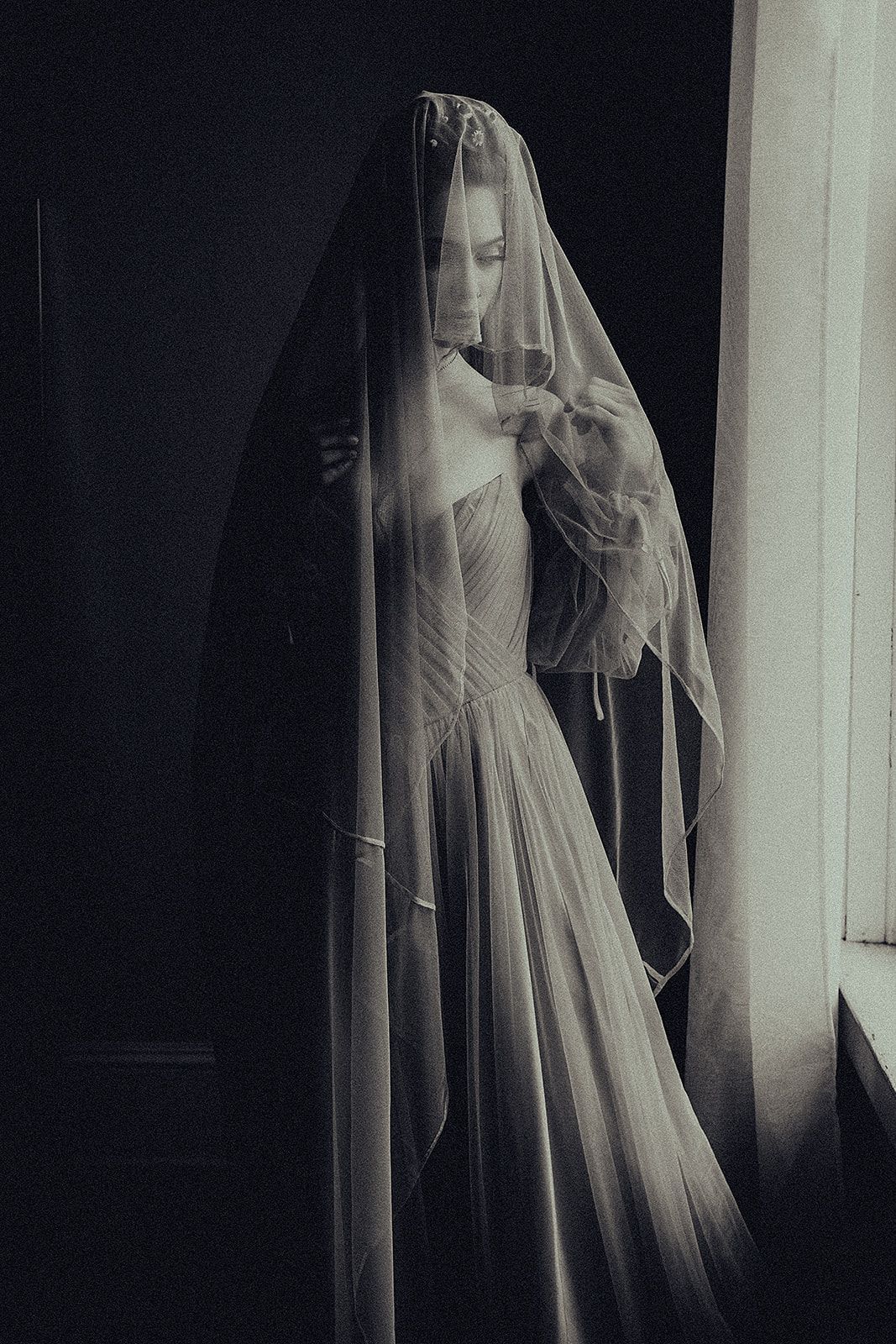 A woman in a wedding dress and veil is standing in front of a window.