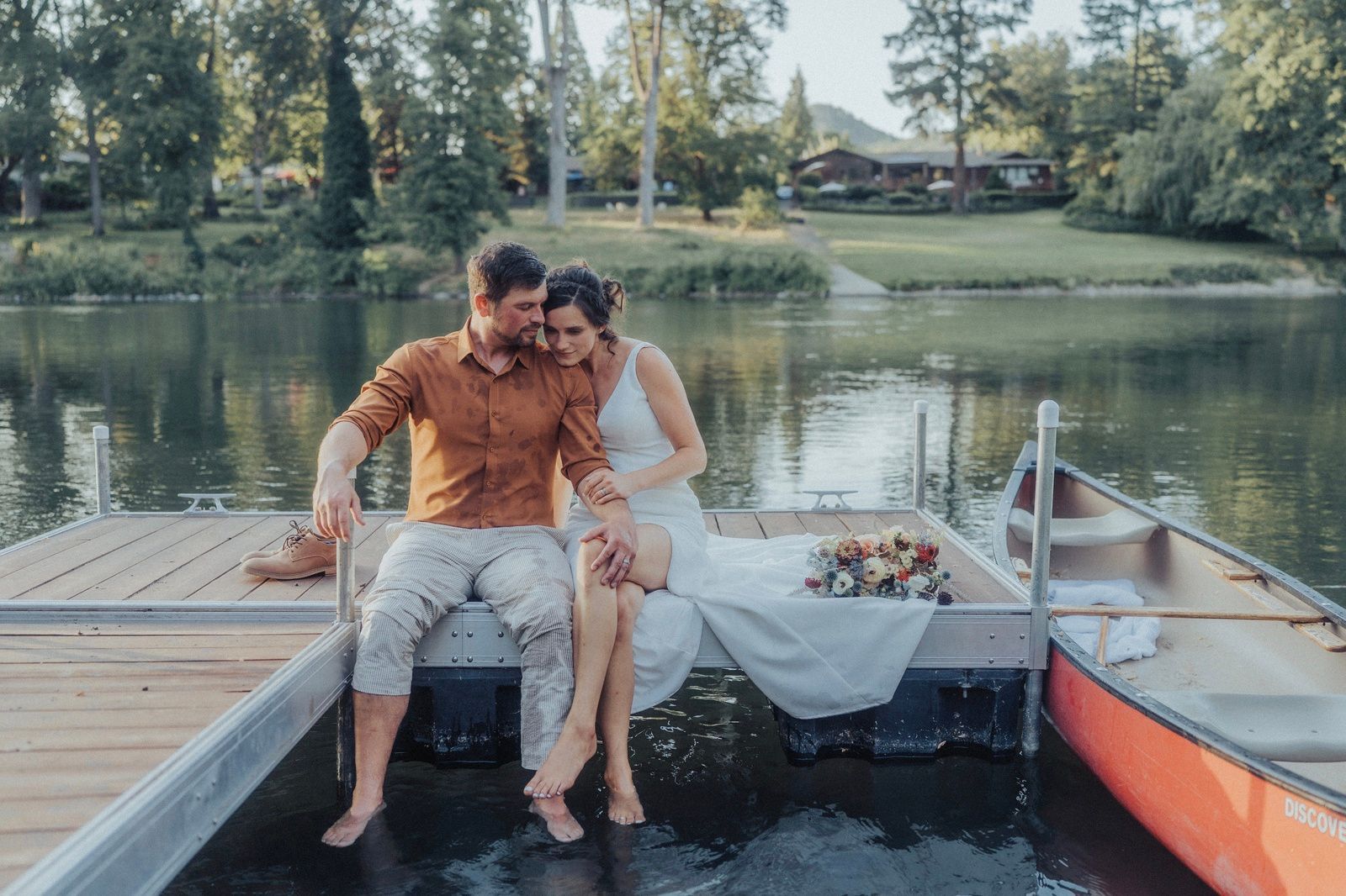 Couple sitting on a dock with bare feet; lake, trees, and canoe visible.