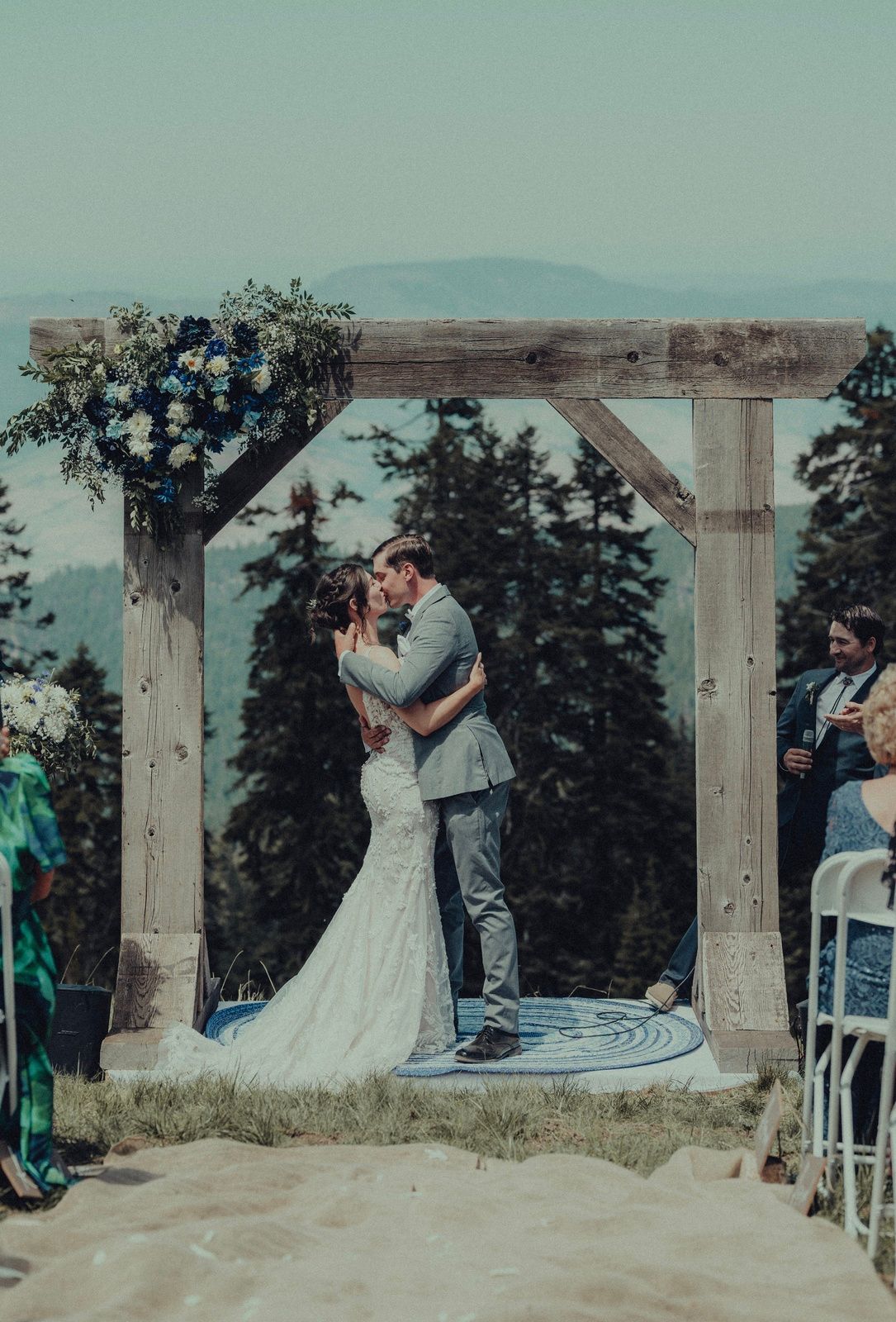 Bride and groom embrace, kissing, under a wooden arch, outdoors with mountain views.