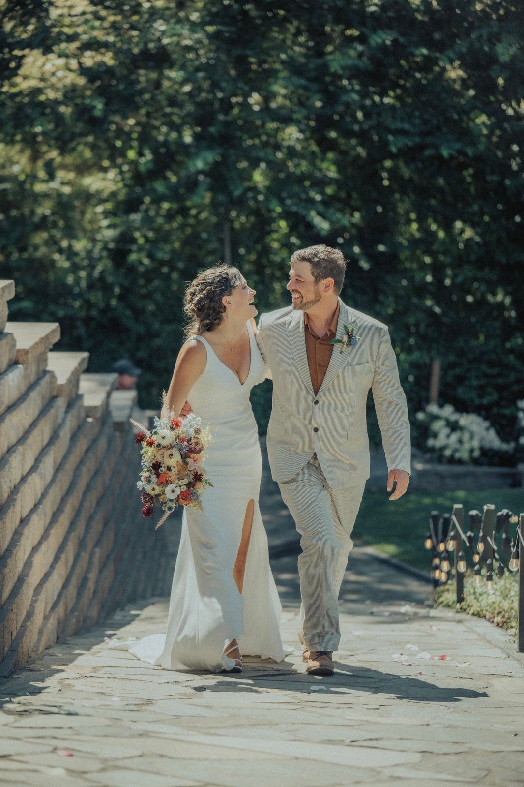 Newlyweds walk arm in arm, smiling, down a stone path. Bride in white dress, groom in beige suit, sunny outdoor setting.