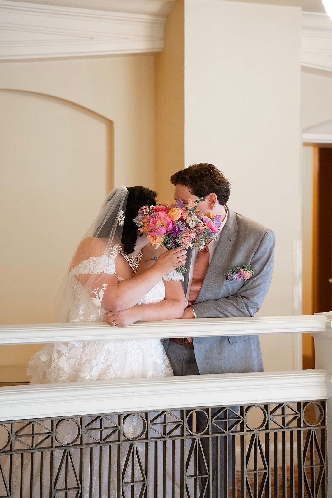 Bride and groom kissing, flowers in between. Bride in white dress, groom in gray suit. Balcony setting.