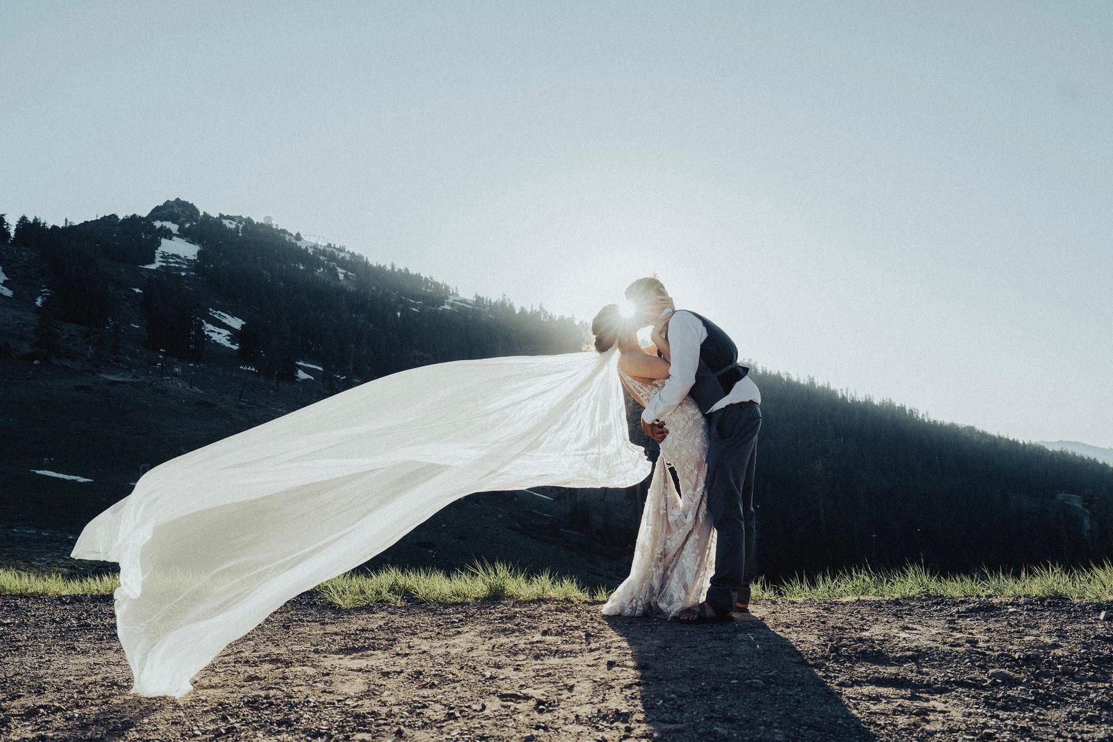 Couple embracing, veil blowing in wind against mountains and sun.