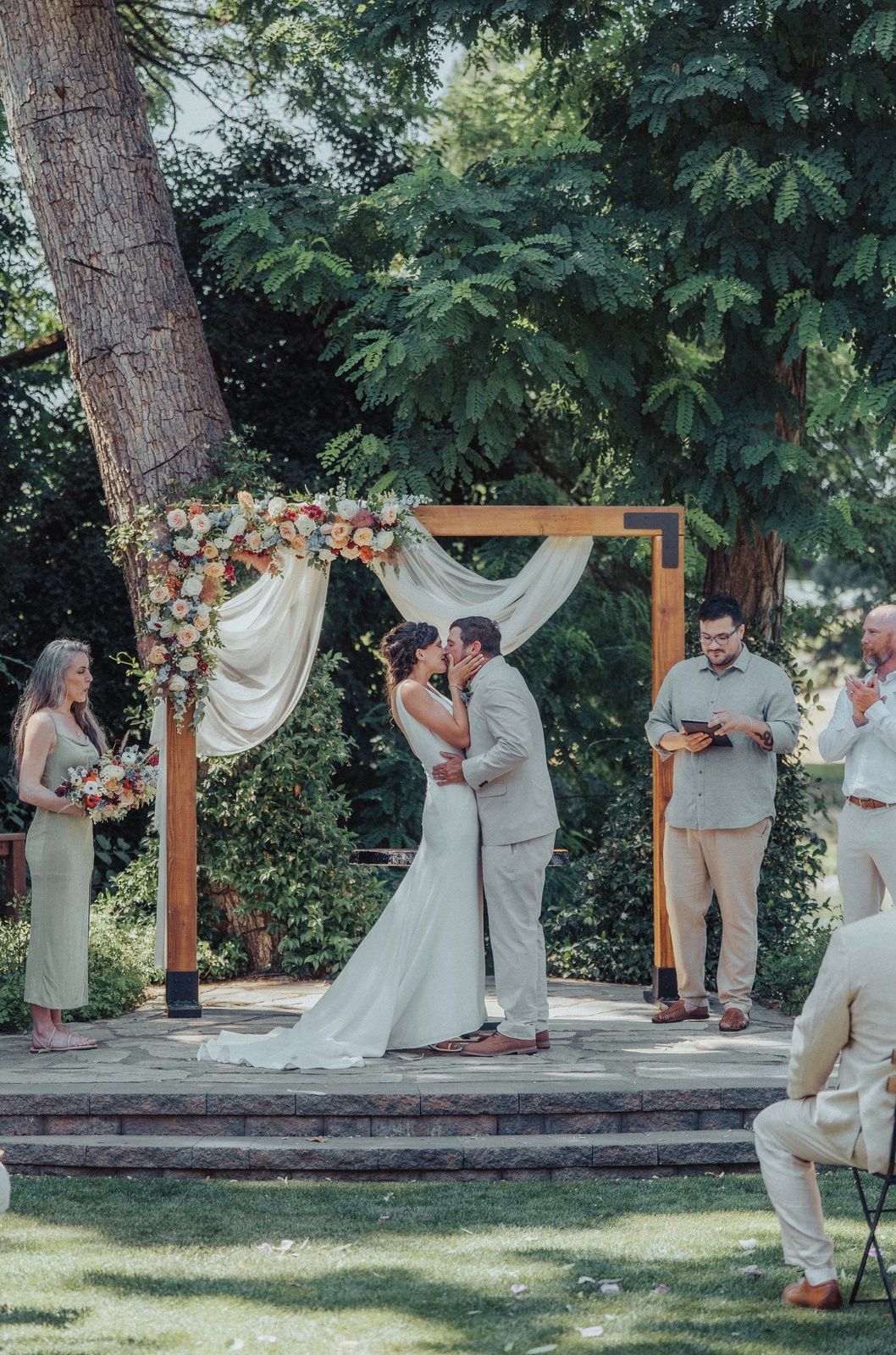 Bride and groom kiss under a floral arch during an outdoor wedding ceremony.