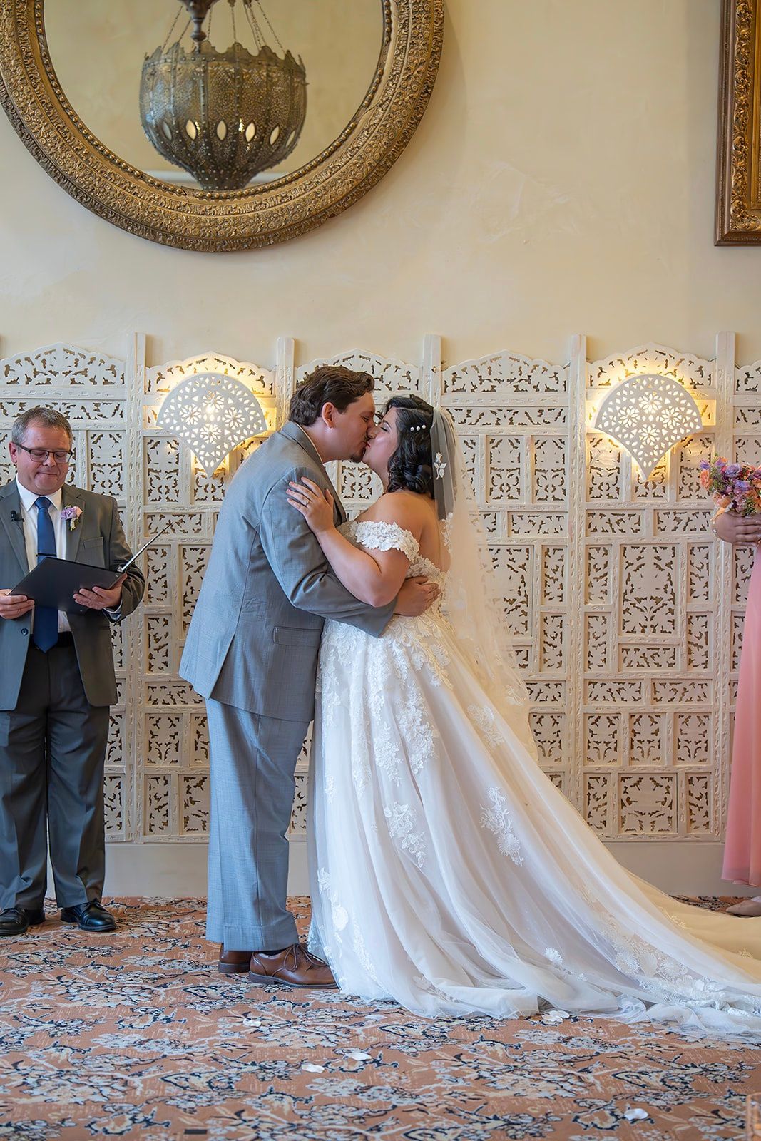 Bride and groom kissing at their wedding ceremony in front of a patterned screen, officiant looking on.