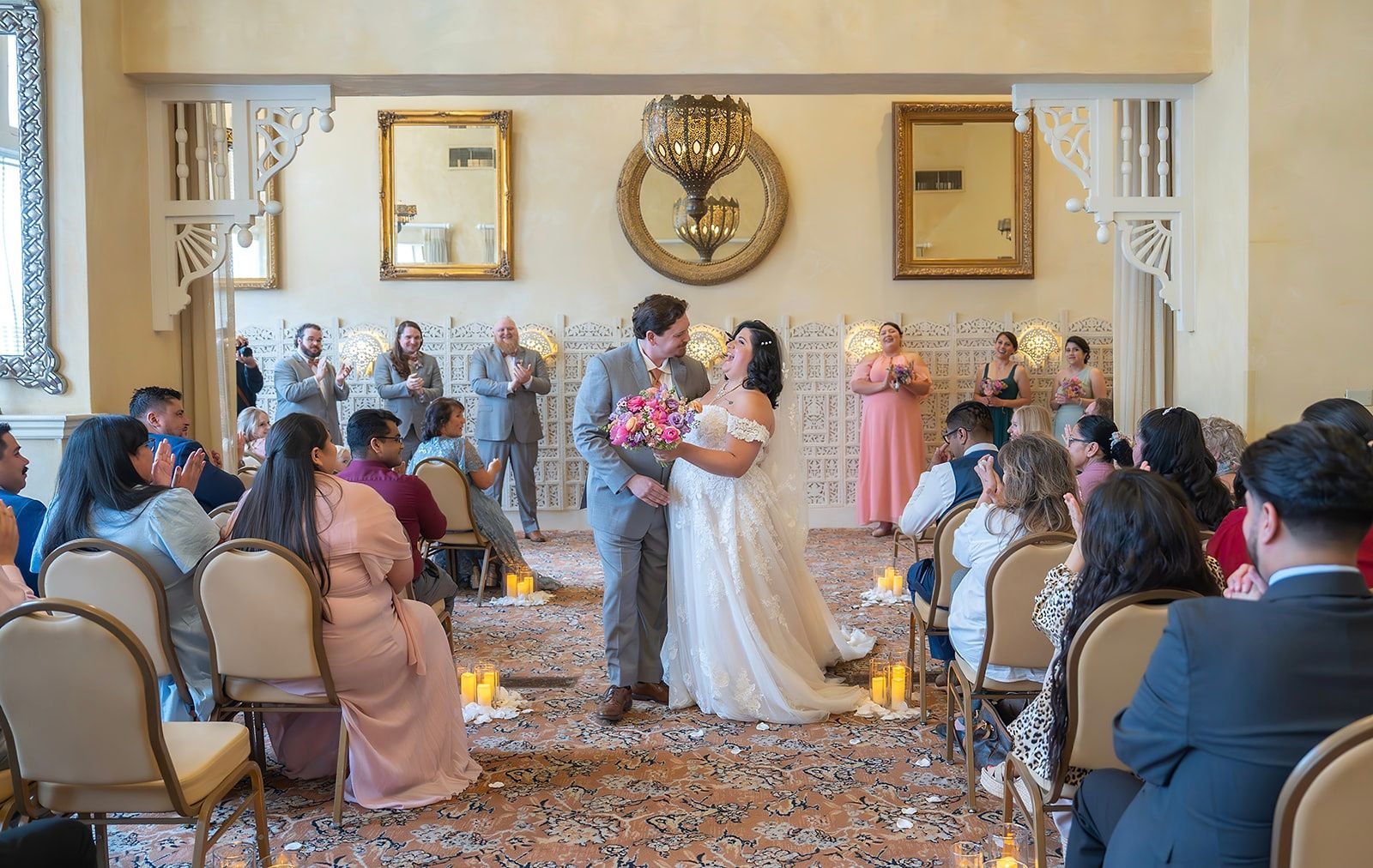 Bride and groom embrace at altar; guests clap. Formal indoor wedding with floral décor and neutral color palette.