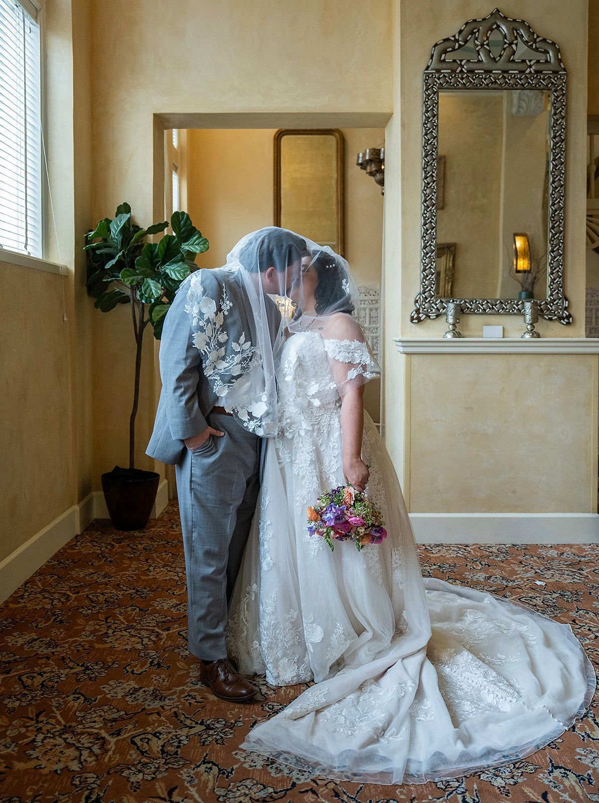 Newlyweds share a kiss, veiled by the bride's veil. They stand in a warmly lit room with a potted plant and ornate mirror.
