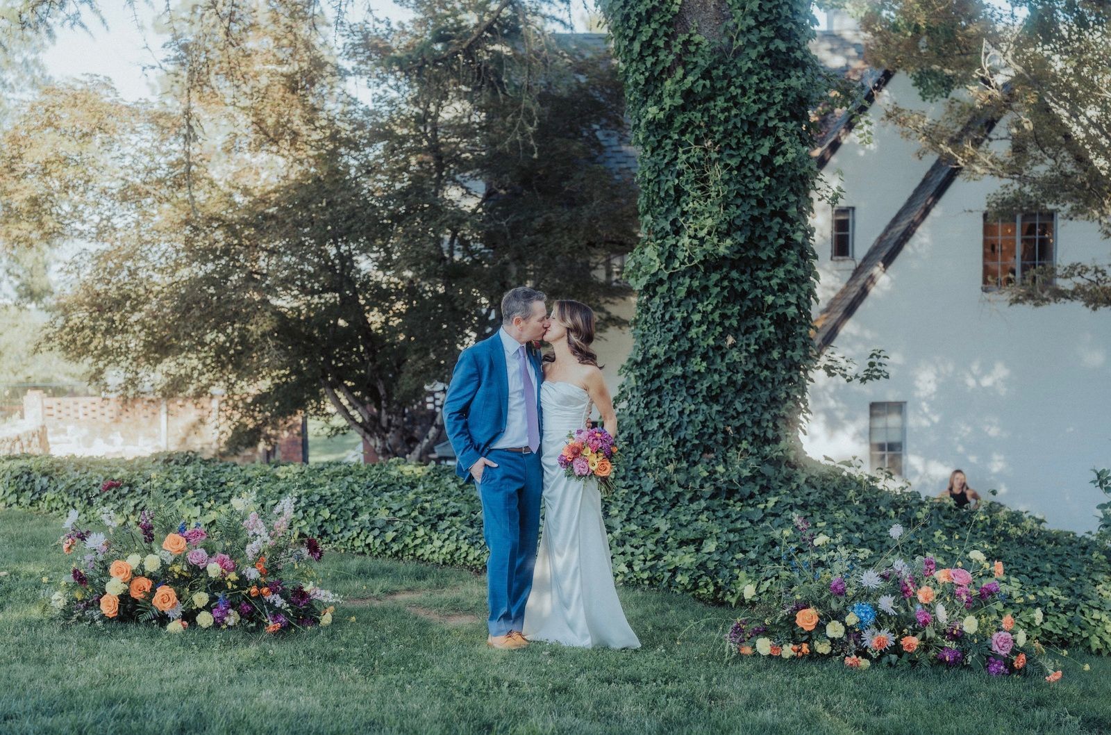 Couple kissing at outdoor wedding ceremony, lush floral arrangements in front of ivy-covered building.