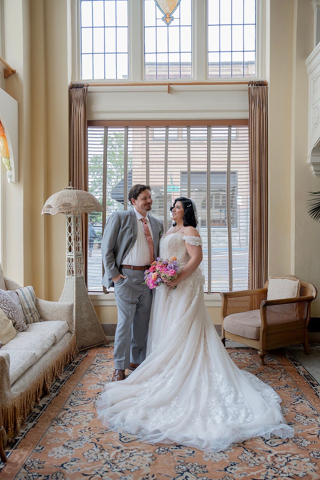A newly married couple stands together in an ornate interior, smiling at each other.