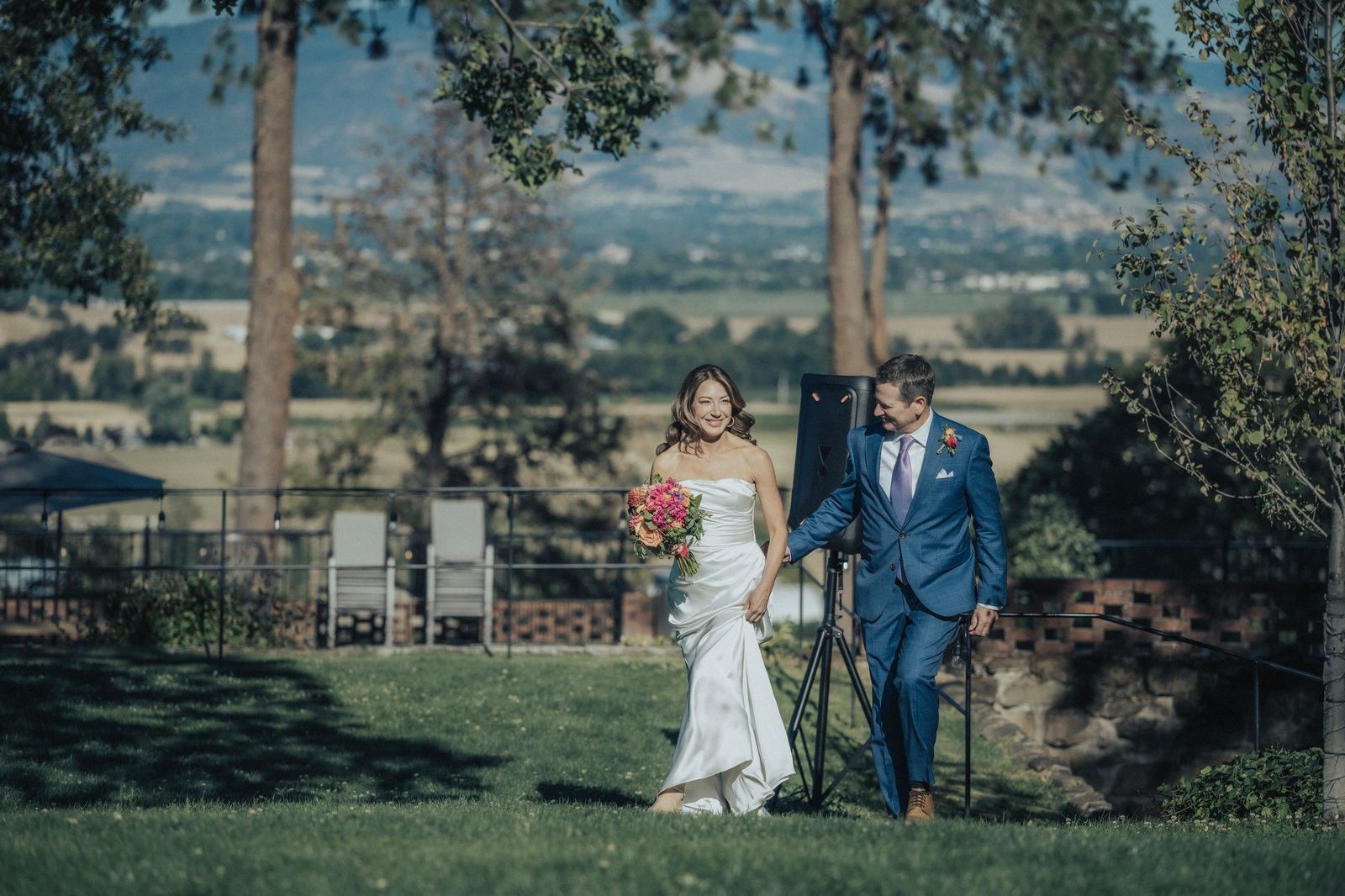 Newlyweds walk hand-in-hand on grass; bride in white gown, groom in blue suit, outdoor setting.