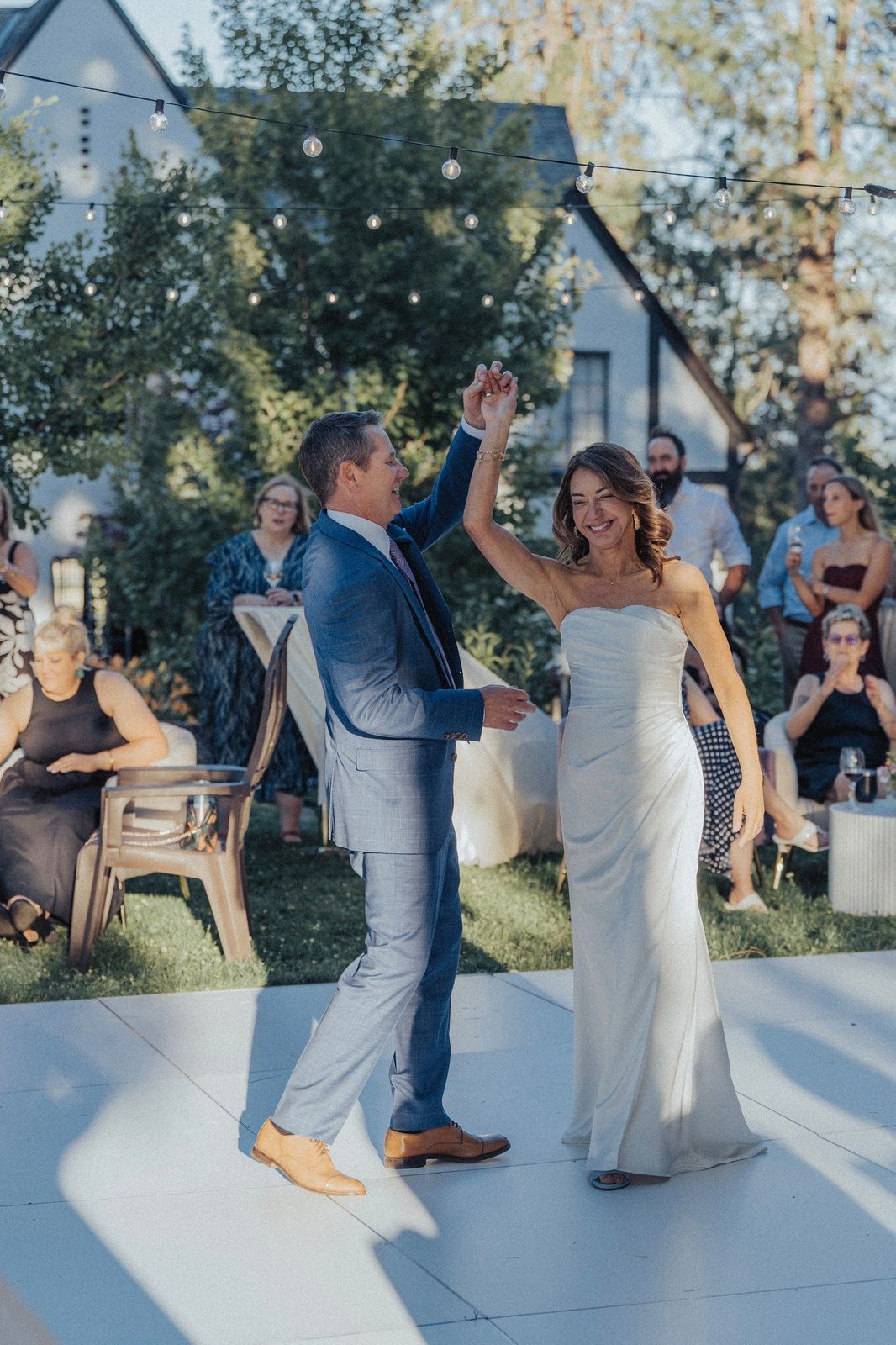 Couple dancing outside, woman in white gown, man in blue suit. Guests watch in the background.