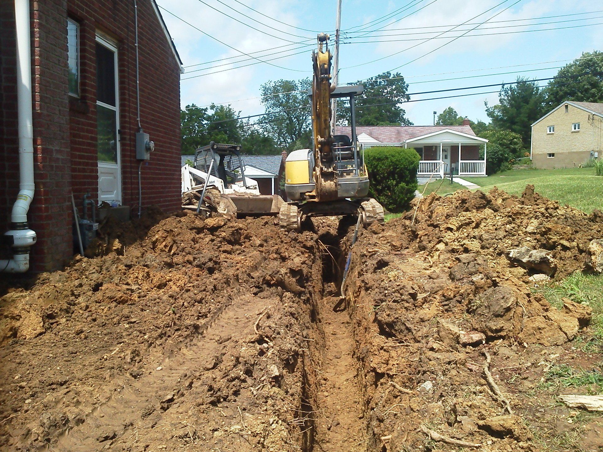 An excavator is digging a trench in the dirt in front of a brick building