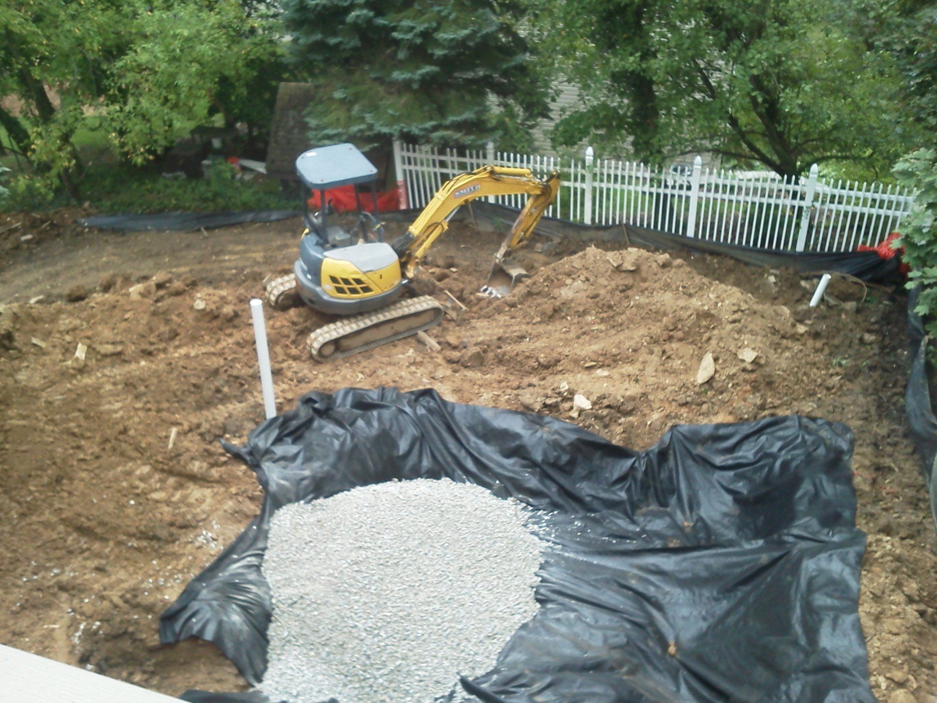 An excavator is digging a hole in the dirt in front of a white fence.