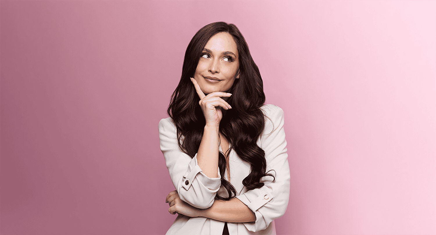 Une femme aux longs cheveux bruns pose devant un fond rose dégradé, la main au menton, le regard détourné.