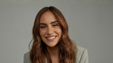 Une personne souriante aux cheveux bruns ondulés, portant un blazer vert clair sur un fond gris uni.