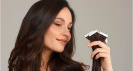 Une personne souriante aux longs cheveux bruns ondulés tient une mèche d'extensions capillaires foncées à bandes adhésives sur un fond gris.