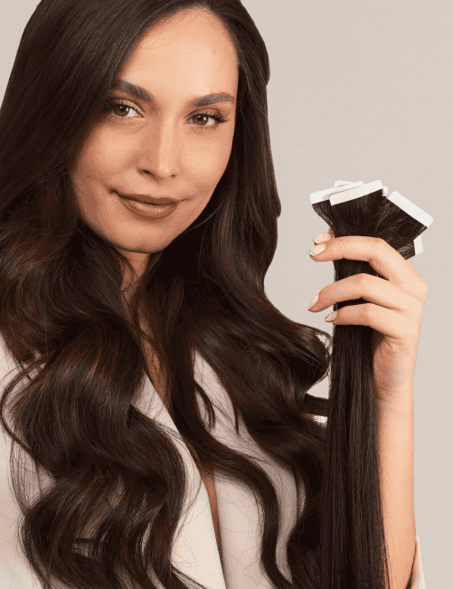 Une personne aux longs cheveux ondulés brun foncé tient à la main un ensemble d'extensions capillaires à bandes adhésives sur un fond uni.