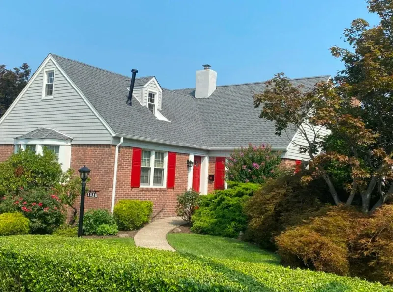A brick and grey-sided house with bright red shutters, a chimney, and a landscaped yard under a clear blue sky.