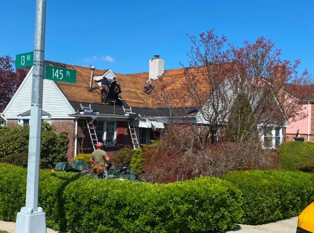Workers replace shingles on a residential house roof, partially covered in new underlayment, near a street sign.