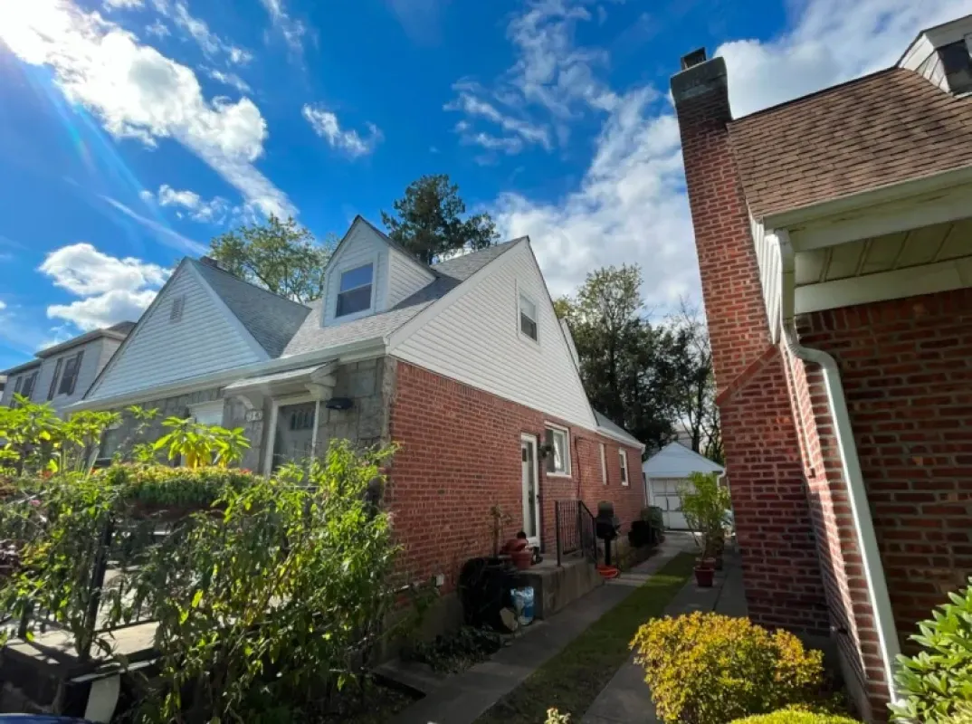 A brick residential home with a white gabled roof and dormer windows, seen from a narrow side yard under a sunny blue sky.