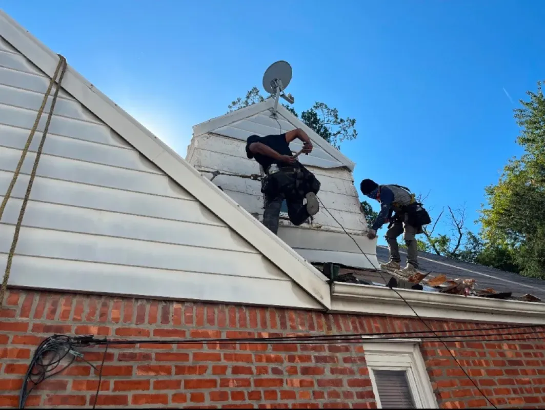 Two workers in safety harnesses perform repairs on the roof of a brick house with white siding under a clear blue sky.
