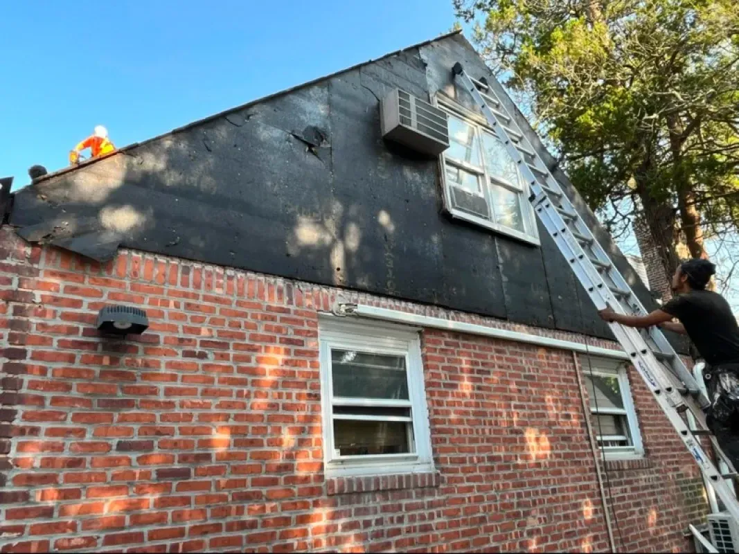 A worker on a ladder performs exterior home repairs on a brick house with black roofing felt on the upper gable.
