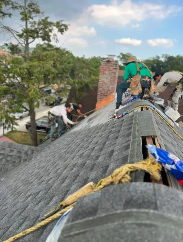 Roofers in safety gear work on a residential roof, installing shingles near a brick chimney on a sunny day.