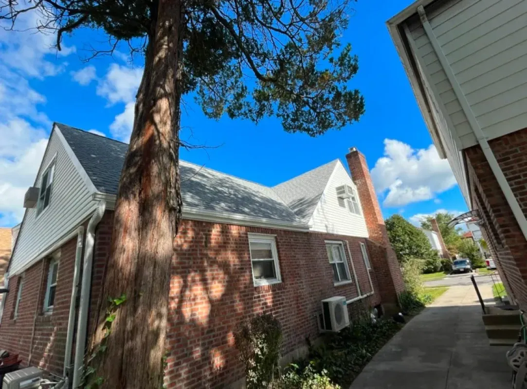 A two-story brick and white siding house seen from a low angle, next to a large tree and a neighboring building.
