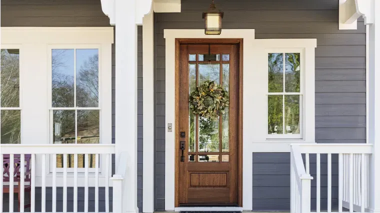 A dark grey house exterior with white trim, featuring a stained wood front door with glass panels and a decorative wreath.