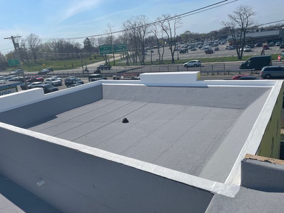 A flat gray rooftop with white parapet walls, surrounded by a parking lot and road in an outdoor, sunny setting.