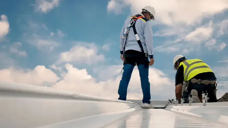 Two workers in hard hats and safety gear installing equipment on a white roof under a bright, cloudy sky.