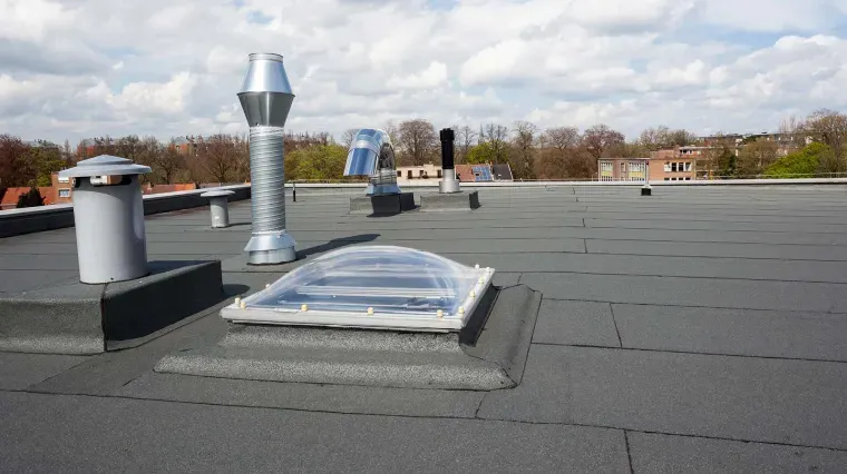 A flat rooftop with dark, textured waterproof membrane, a clear skylight dome, and various metal ventilation pipes.