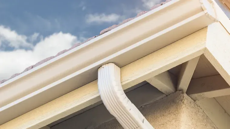 A white metal gutter and downspout attached to the eaves of a house against a blue, cloudy sky.
