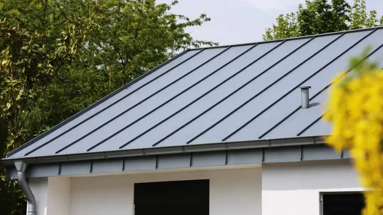 A metal standing-seam roof on a white house, framed by green trees and a blurred yellow flowering bush.