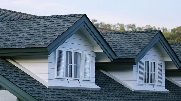 A close-up of a house roof featuring two white dormer windows with shutters against dark gray shingles.