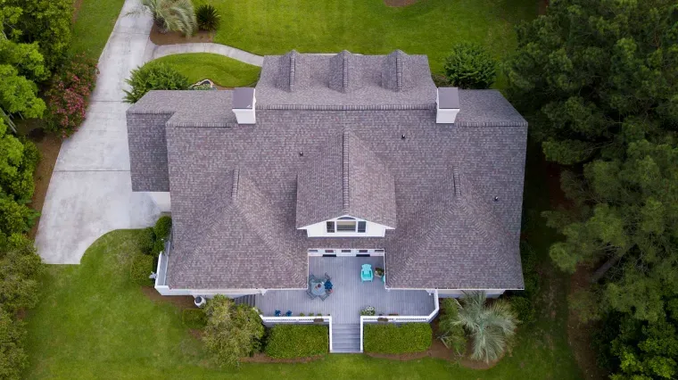 A high-angle aerial view of a house with a gray shingled roof, a back patio, and a driveway surrounded by trees and grass.