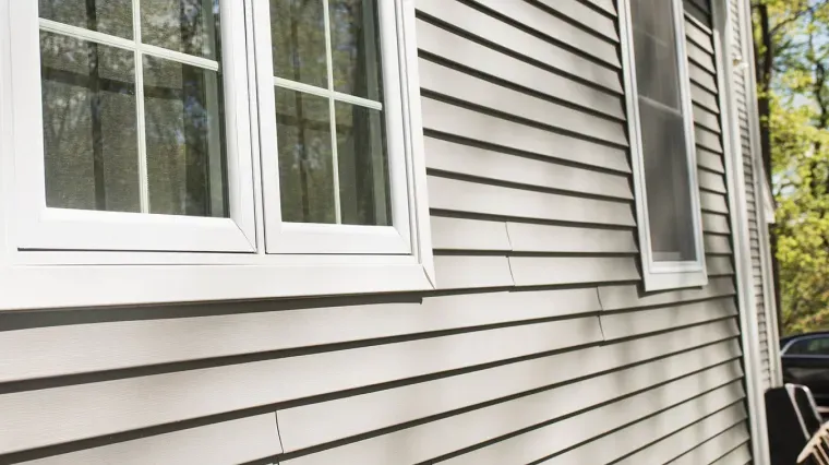 Light gray horizontal vinyl siding on the exterior wall of a house, featuring two white-framed windows.