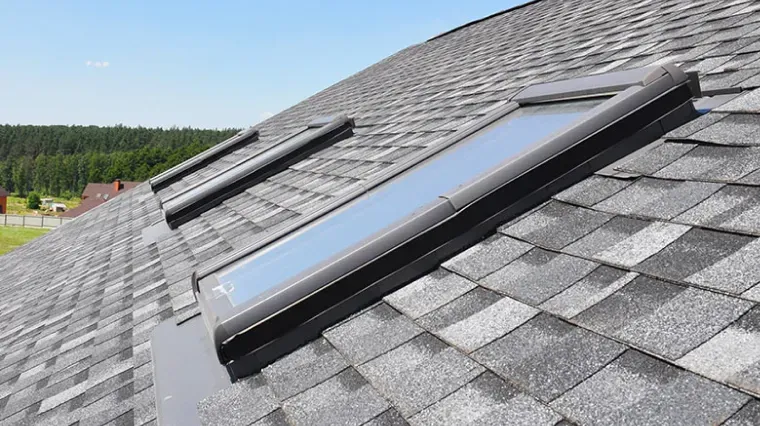 Two rectangular skylights installed on a sloped residential roof covered in gray asphalt shingles against a blue sky.