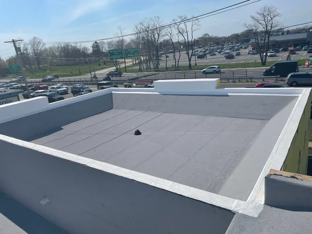 A flat, grey roof with white parapet walls, overlooking a parking lot and a road under a bright, clear sky.