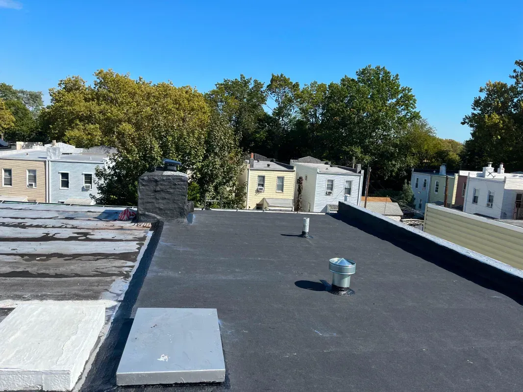 A flat black roof with vents and a chimney, overlooking a neighborhood of row houses and green trees under a blue sky.