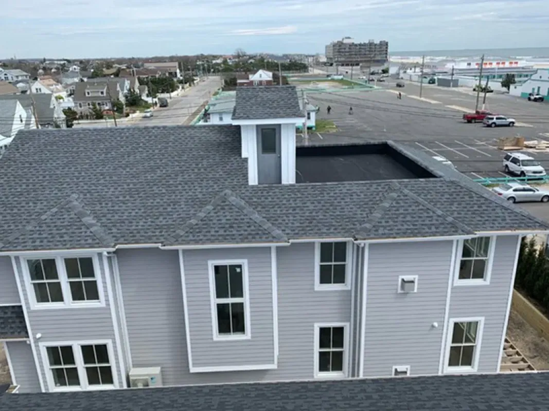 An aerial view of a gray house with a new dark shingled roof and a flat roof section, located in a coastal neighborhood.