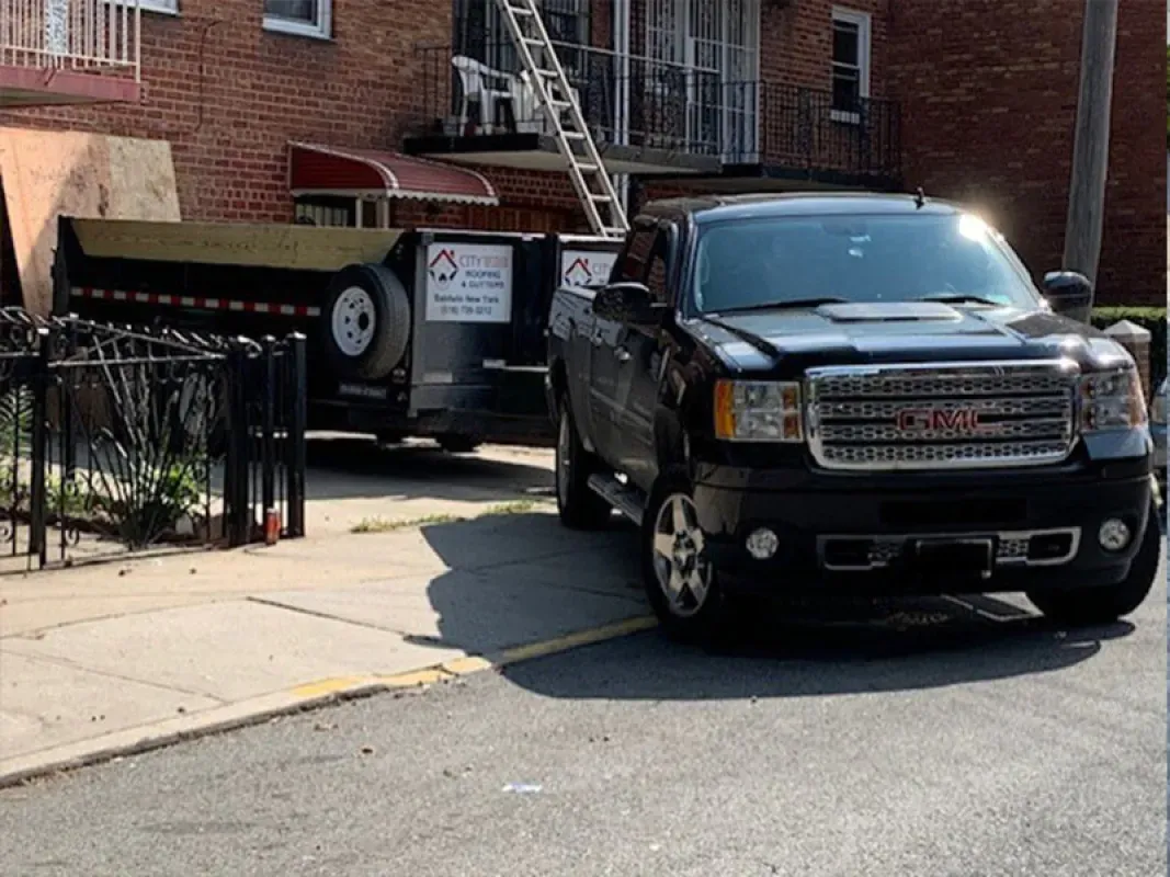 A black GMC truck parked on a street in front of a house, towing a large dumpster with a ladder resting against the building.