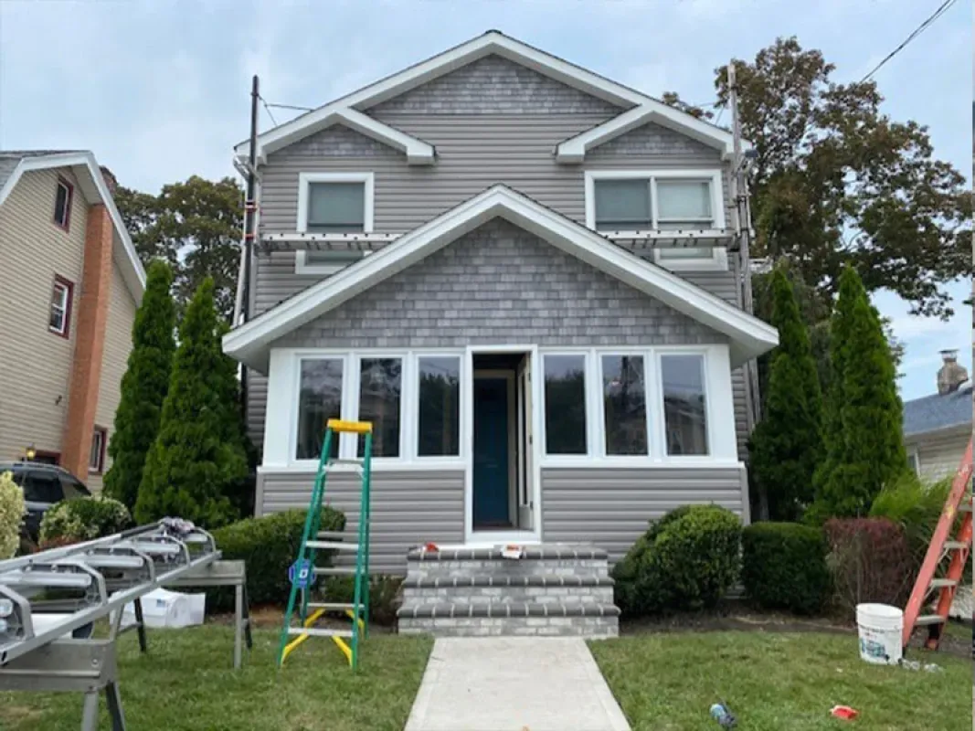 A two-story grey house with a front porch, cedar shake siding, white trim, and a brick staircase under construction.