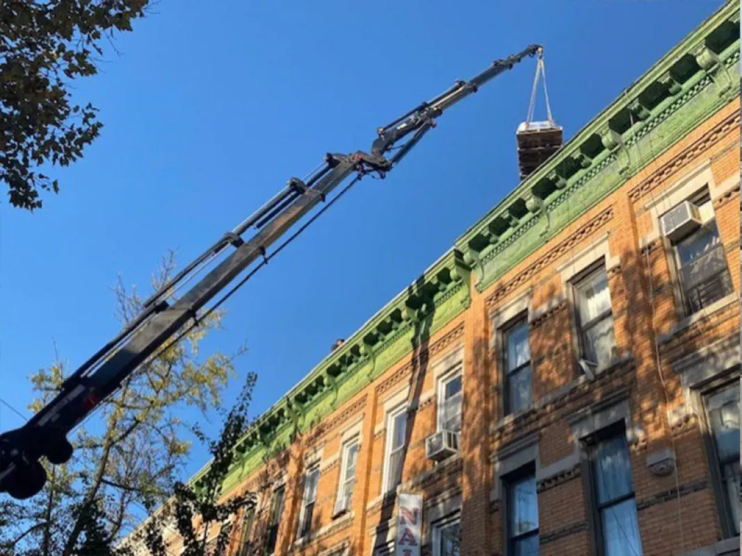 A truck-mounted crane lifts a heavy object toward the roof of a multi-story brick apartment building under a blue sky.