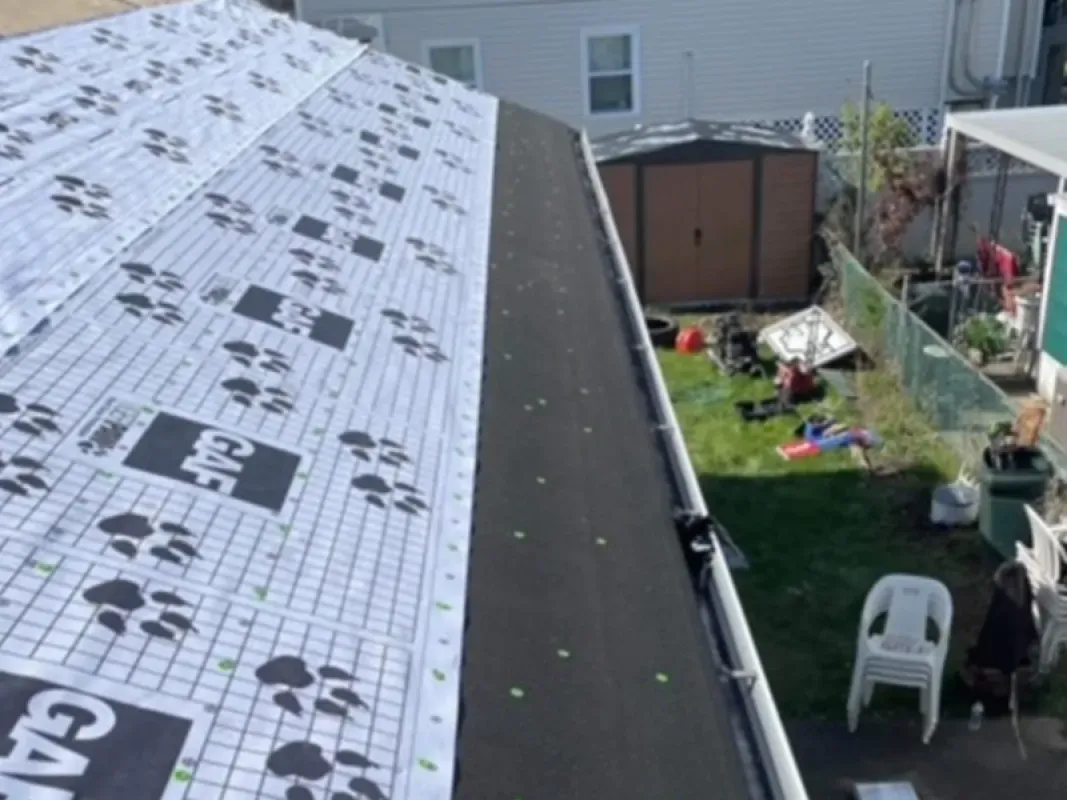 A roof under construction featuring white GAF underlayment, a dark starter strip along the edge, and a view of a backyard.