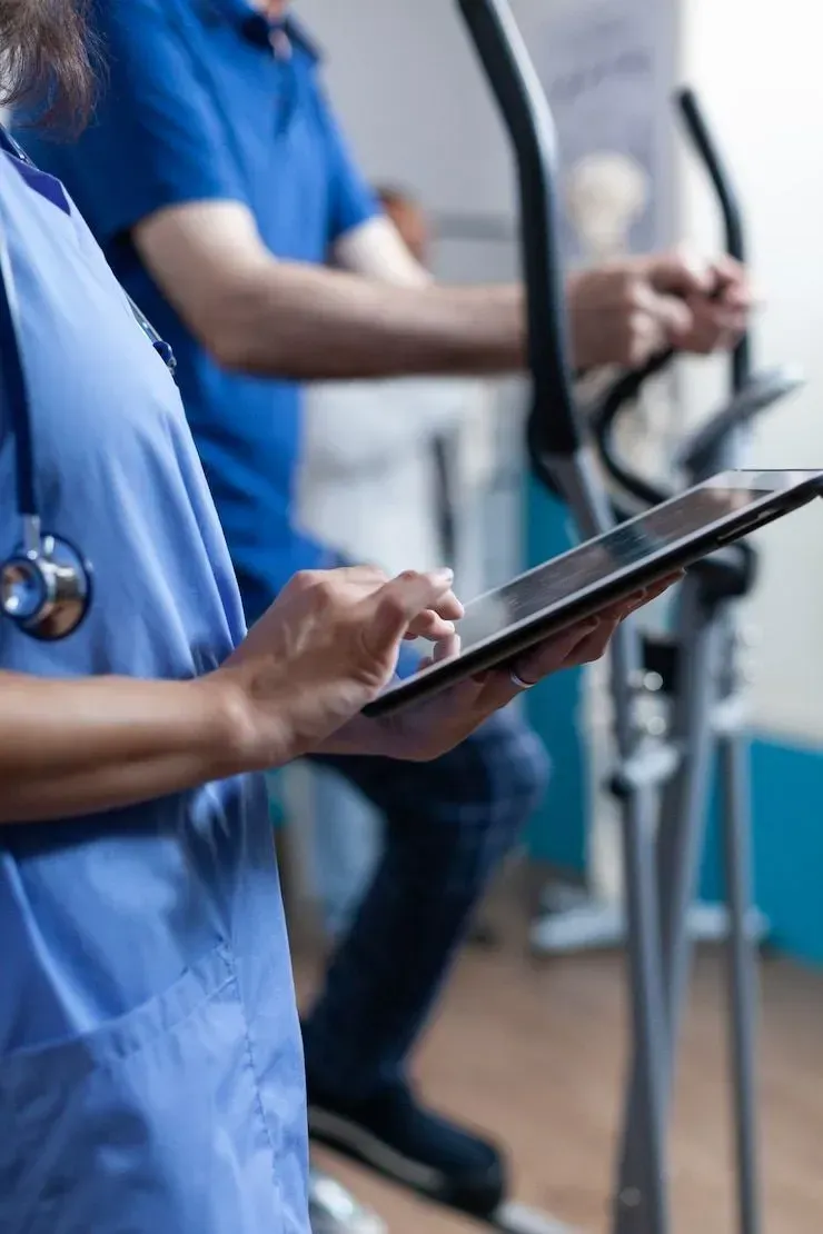 A nurse is using a tablet while a man is riding an elliptical machine.