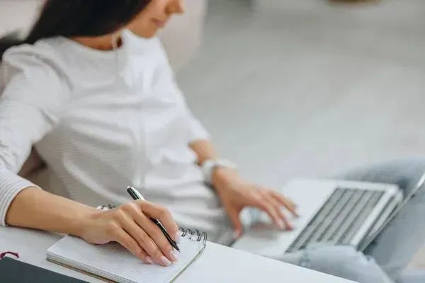 A woman is writing in a notebook while using a laptop computer.